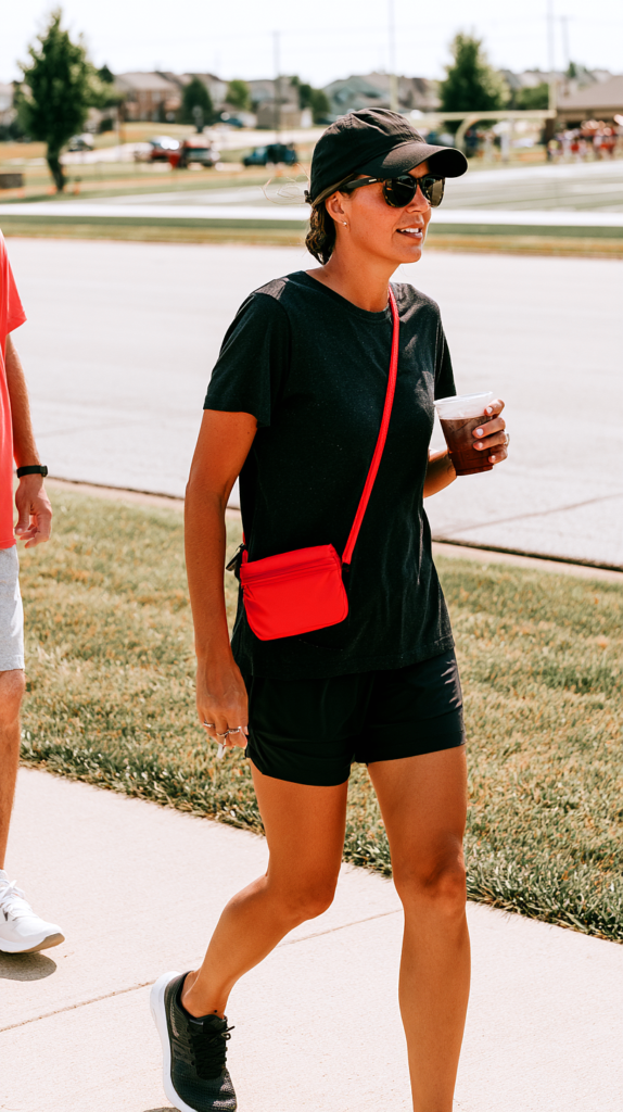 Black tee and athletic shorts sports mom outfit with red crossbody bag and baseball cap