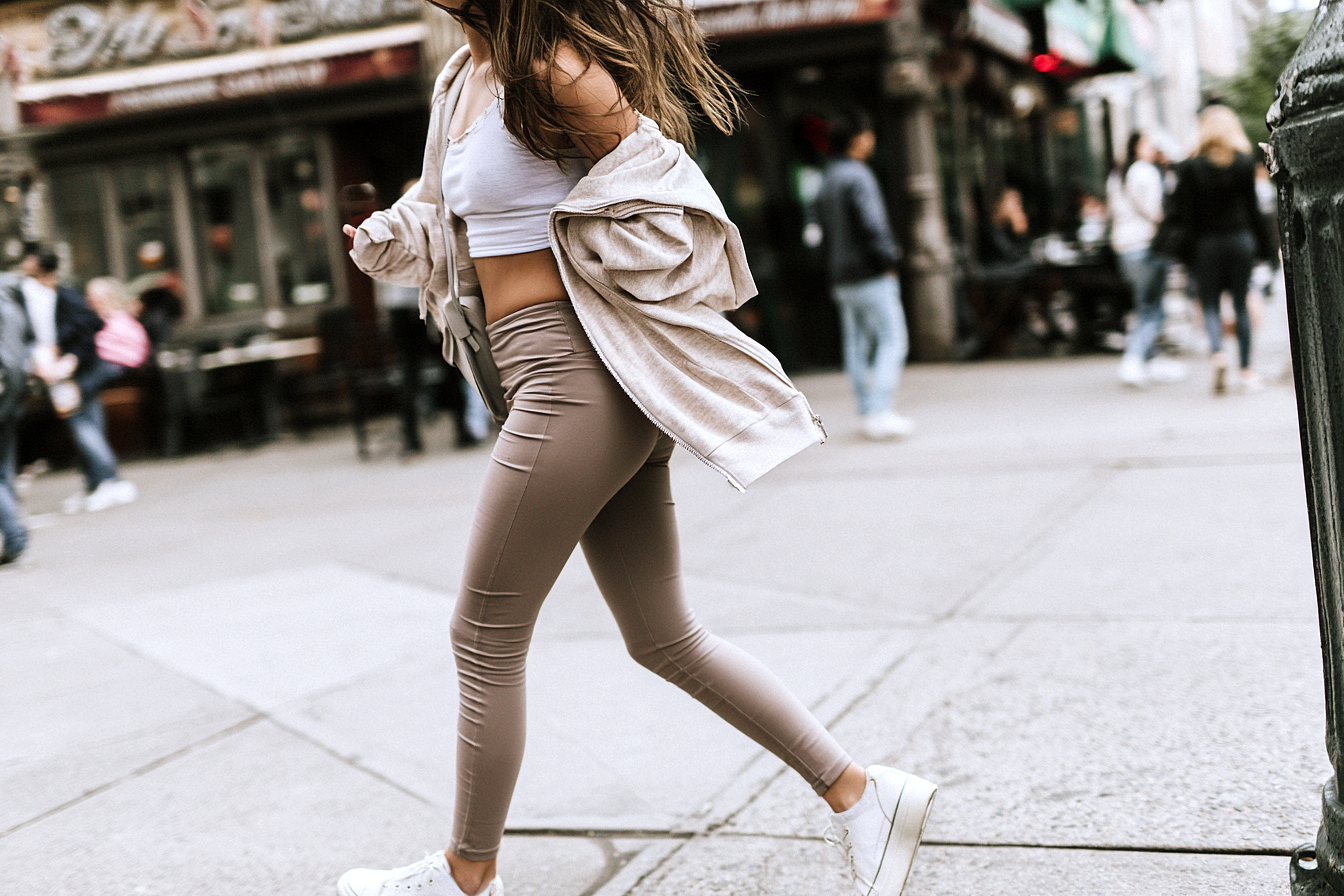 Woman walking on a city sidewalk in taupe leggings white crop top and sneakers  sporty-casual street style outfit guide